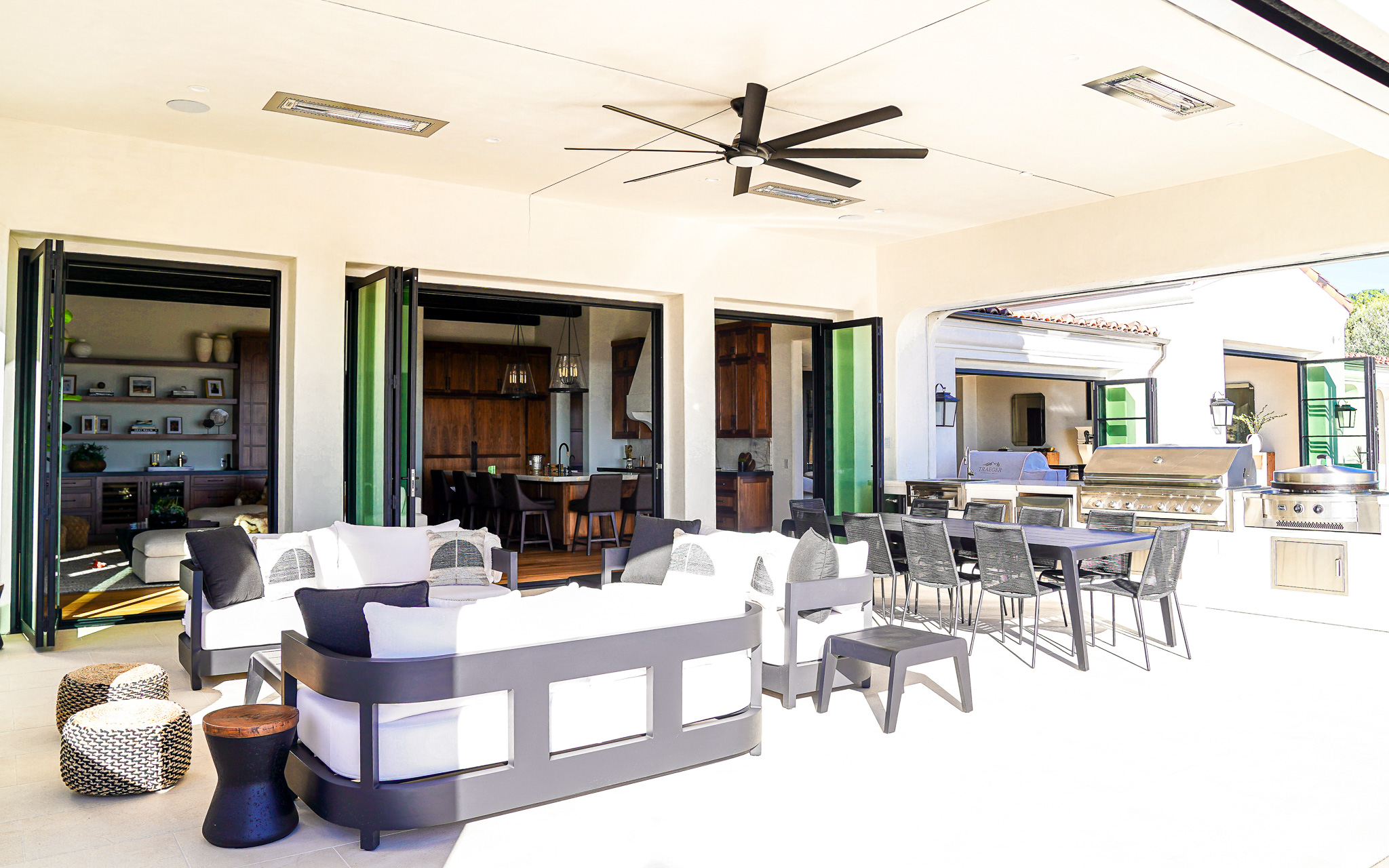Luxurious outdoor patio area with a modern seating arrangement, ceiling fans, open folding doors, and an integrated kitchen with bar stools.