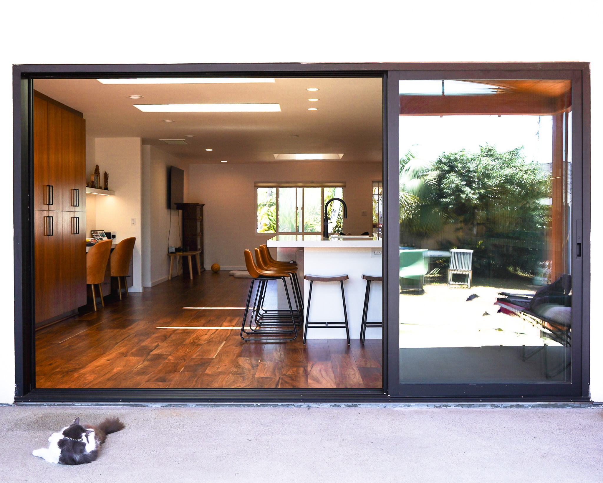 A contemporary kitchen viewed through a large open sliding glass door, with a cat lying on the ground in the foreground.