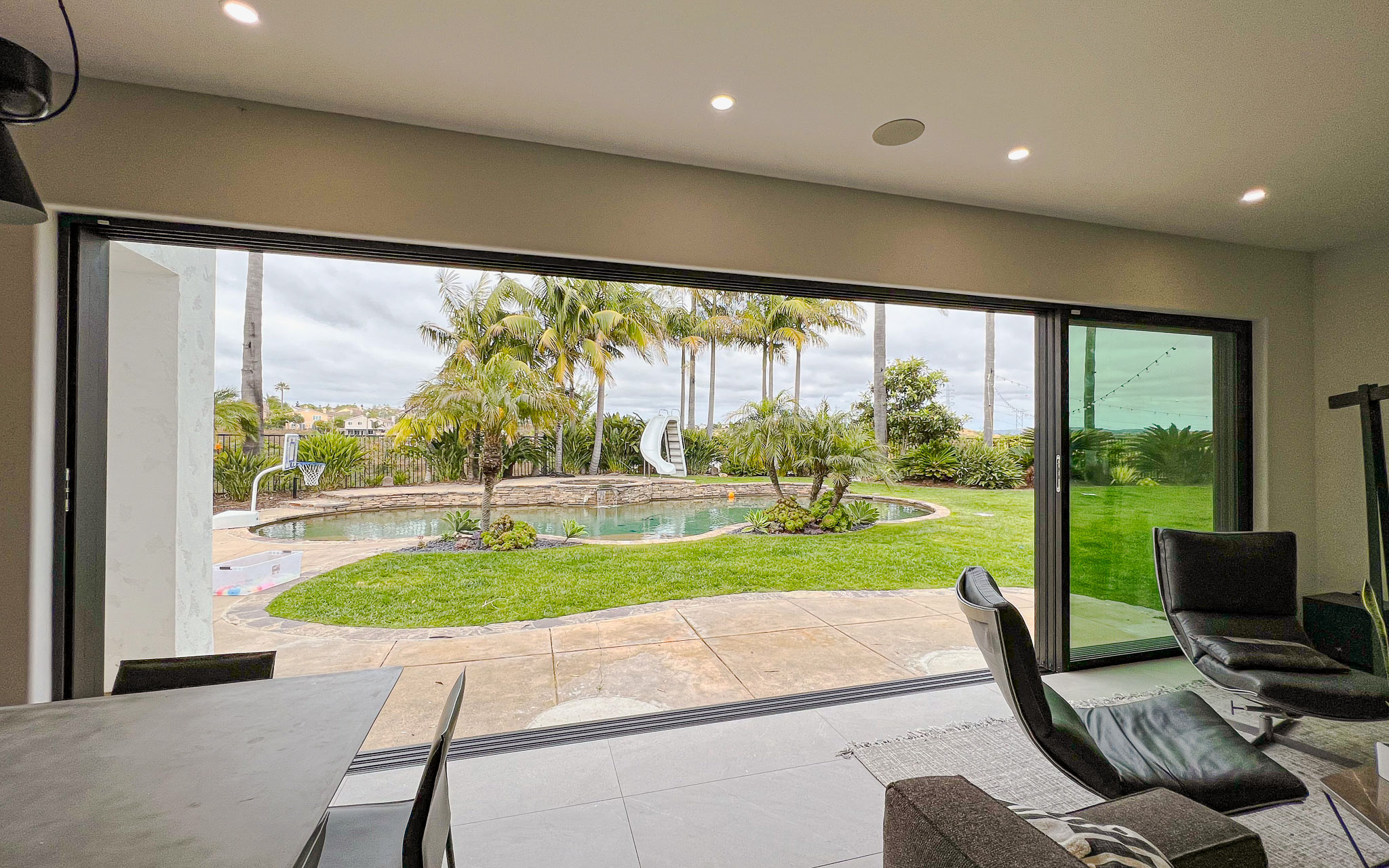 Modern living room with large sliding glass door overlooking a backyard with a swimming pool and tropical landscaping.