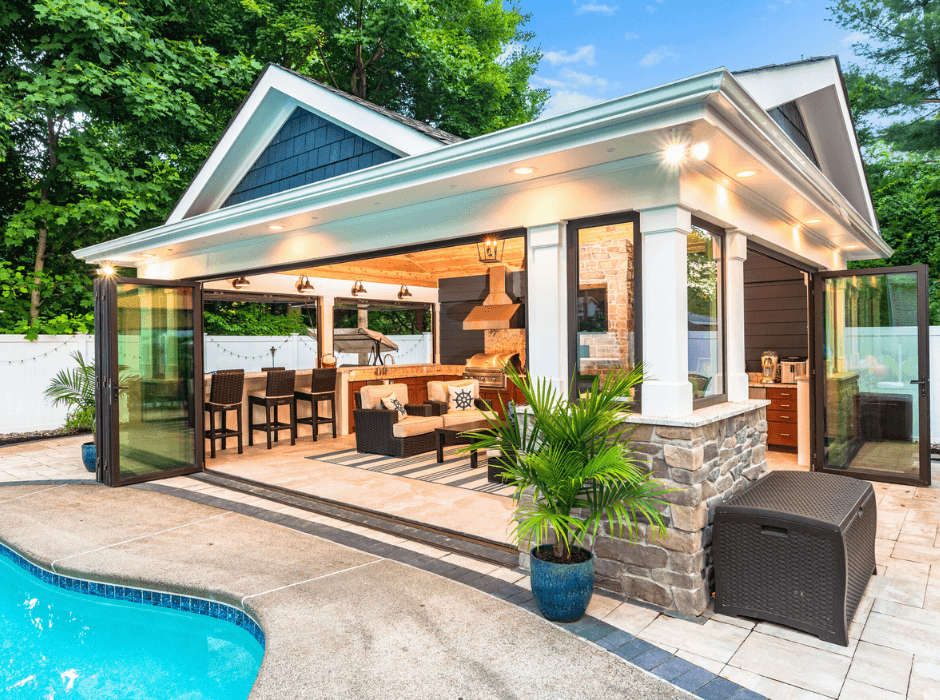 Outdoor living space with a pool, featuring a covered area with an open kitchen, bar seating, a couch, plants, and sliding glass doors. The area is surrounded by greenery and a white fence.