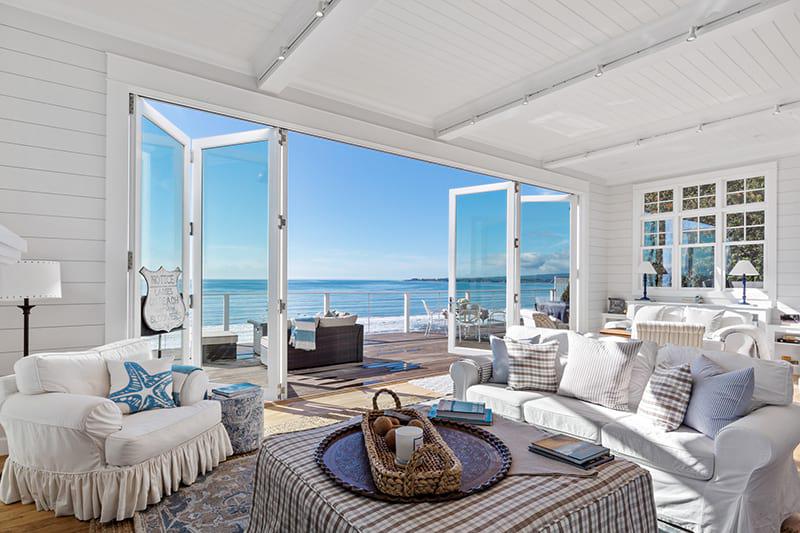 Spacious beach house living room with white furniture, light blue accents, and large open glass doors leading to a wooden deck overlooking the ocean.