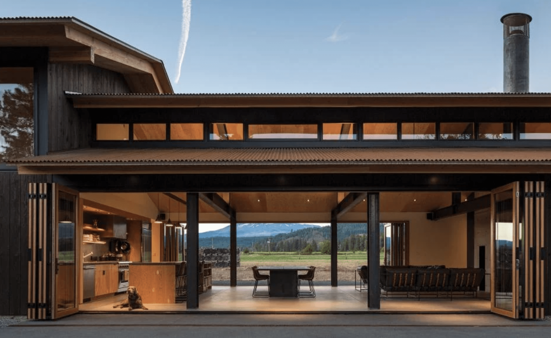 Contemporary house with large sliding glass doors open to an outdoor space. The interior showcases a kitchen and seating area. A view of a landscape with mountains is visible through the doors.