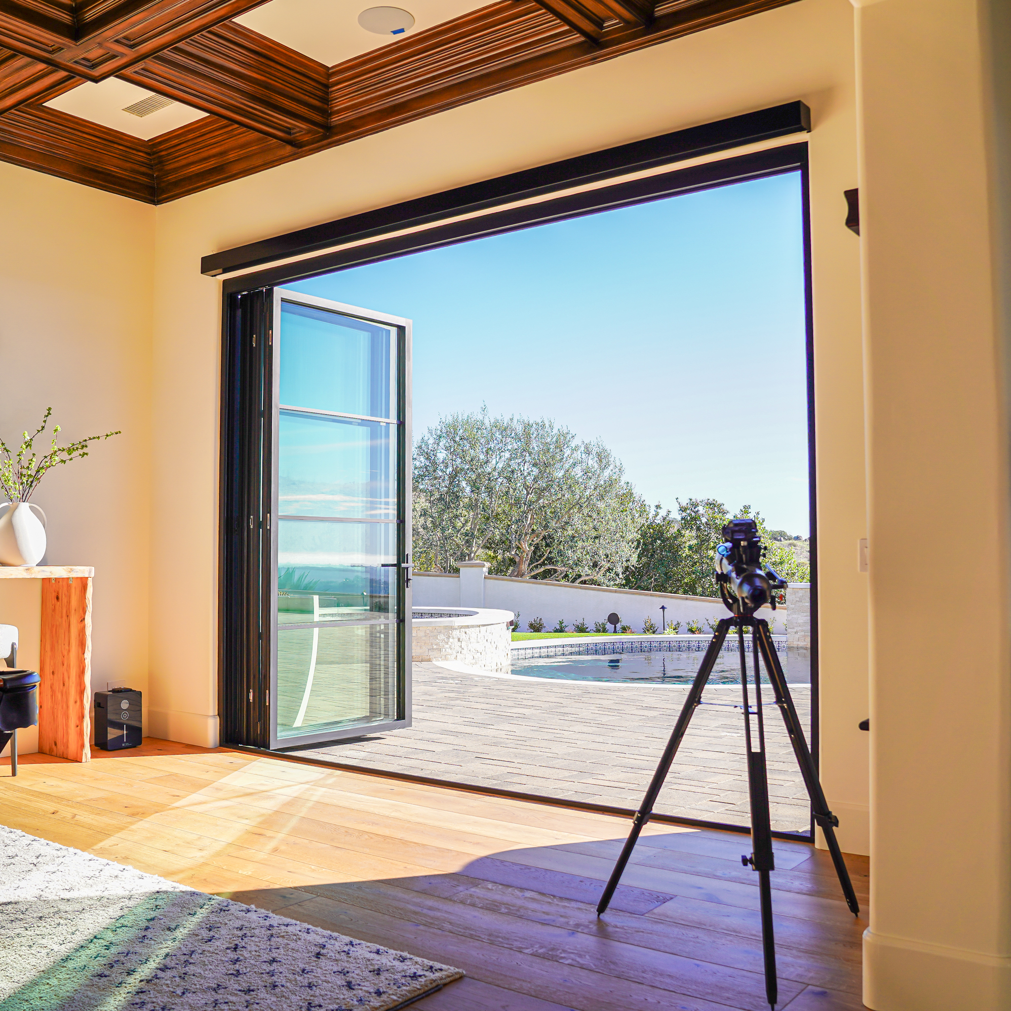 A camera on a tripod is set up at an open patio door, overlooking a terrace with a pool and trees beyond, shot from inside a modern room with wooden floors and ceiling.