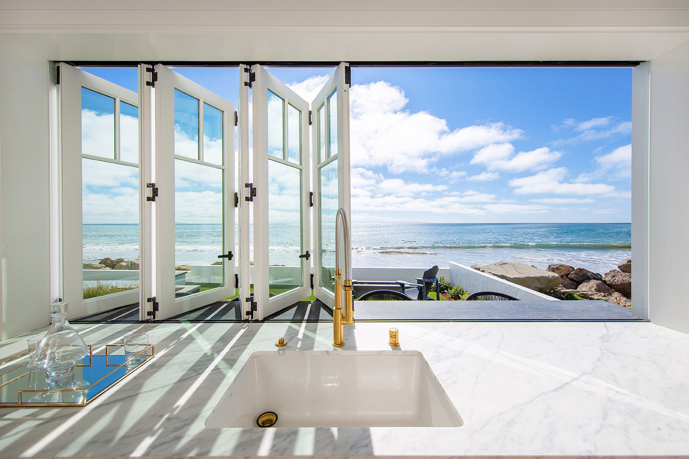 A kitchen sink with a marble countertop is positioned in front of open windows offering a clear view of a sunny beach and ocean.
