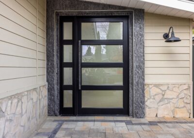 Modern front door with frosted glass panels and black frame surrounded by light-colored siding and stone veneer. A wall-mounted outdoor light fixture is visible on the right side.