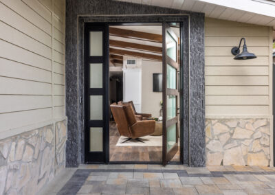 A modern entry door leads to a living room with two brown leather chairs, stone tile flooring, and a wall-mounted light fixture outside.