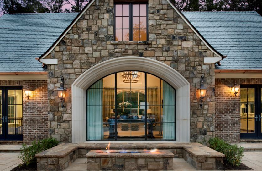 A stone and brick house with a large arched window, elegant lighting fixtures, and a modern fire feature in the foreground.