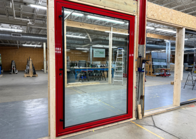 A large window with a red frame installed in an unfinished wall inside a warehouse. Vinyl siding, ladders, and construction materials are visible in the background.