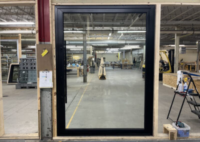 A large glass door with a black frame installed in an industrial space. The background shows various workshop equipment and materials.