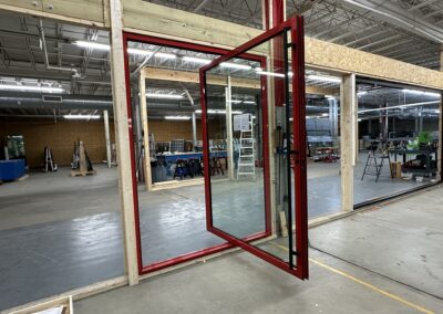 A red-framed glass door is partially open in a large warehouse space, with various tools and ladders scattered around. Wooden framing lines partial walls.
