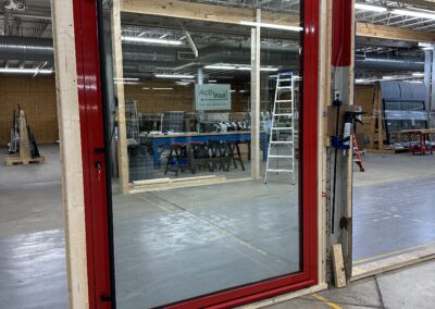 A large red-framed glass door installed in a wooden frame inside a warehouse, with industrial equipment and ladders visible in the background.