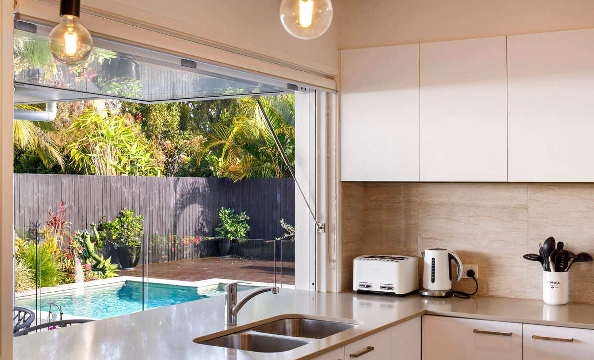A modern kitchen with white cabinets and a sink overlooks a backyard with a swimming pool. An electric kettle and a toaster are on the counter. The backyard features lush greenery and a wooden fence.