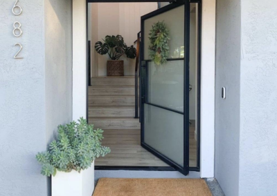 Front entrance of a modern home with gray walls, a large glass door, and house number 682. The design features a succulent planter and a doormat. Inside, stairs lead to the main living area with green plants.