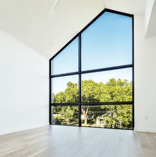 A room with light wood floors and white walls features large, triangular floor-to-ceiling windows overlooking green trees and a clear blue sky.